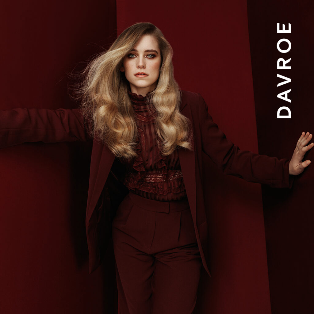 Kobieta o długich, gęstych i pofalowanych włosach w odcieniu miodowego blondu, modelka marki Davroe w bordowej stylizacji.